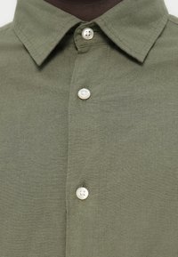 Πουκάμισο σε ελιά πράσινο με κλασικό γιακά, διαθέτει εμπρόσθιο καβάλο με λευκά κουμπιά και απαλό, υφασμάτινο ύφος.