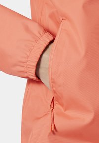 Jasnoperłowa, lekka kurtka w kolorze brzoskwiniowym z elastycznymi mankietami, wyposażona w boczną kieszeń zapinaną na zamek oraz regulowany sznurek dla dopasowania.