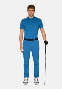 Blå pikétröja med korta ärmar och vita detaljer, matchad med blå byxor och vita sneakers. Håller en golfklubba.