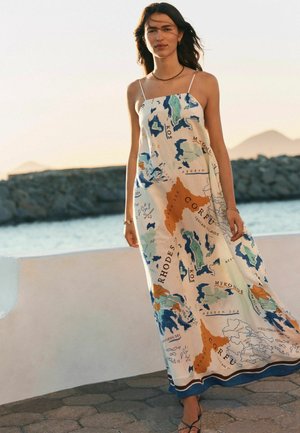 Žena stojí vonku pri vode, oblečená v dlhé, bezrukávové šaty s farebnou mapou gréckych ostrovov a morí.