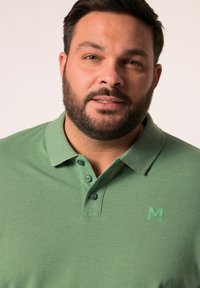 Zielona koszulka polo z teksturowanej tkaniny, trzy guziki przy kołnierzy oraz małe zielone logo "M" na piersi. Zbliżenie.
