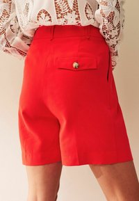 Rode shorts met een gladde textuur, voorzien van een achterzak met knoopsluiting. De tailleband zit net boven de heupen.
