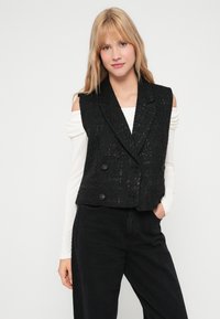 Czarna, teksturowana, dwurzędowa kamizelka z szerokim kołnierzem, guzikami i wycięciami na ramionach, noszona na długim białym topie.