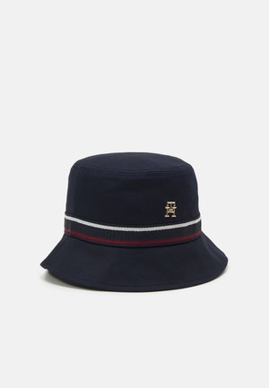 Chapeau bob bleu marine à large bord, avec un bandeau rayé noir, blanc et rouge, et un petit emblème doré sur le côté.