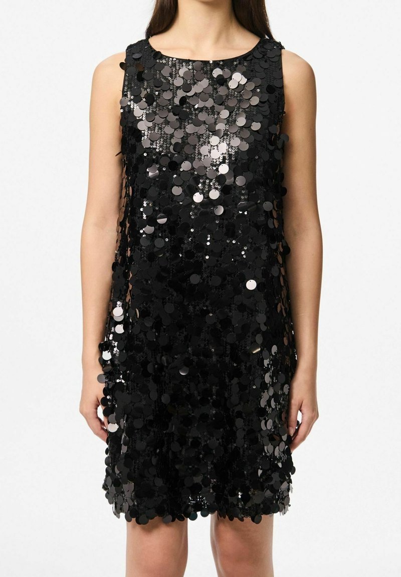 Robe mini noire sans manches recouverte de grands sequins brillants, portée par une personne debout, les bras détendus le long du corps.