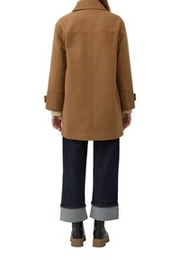 Person in einem braunen Wollmantel, dunklen, gefütterten weit geschnittenen Jeans und schwarzen klobigen Stiefeln, der von hinten auf einem weißen Hintergrund steht.