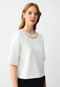 Witte korte mouw top met een ronde halslijn, voorzien van een decoratieve kristallen versiering langs de kraag, gemaakt van zachte, textuurstof.