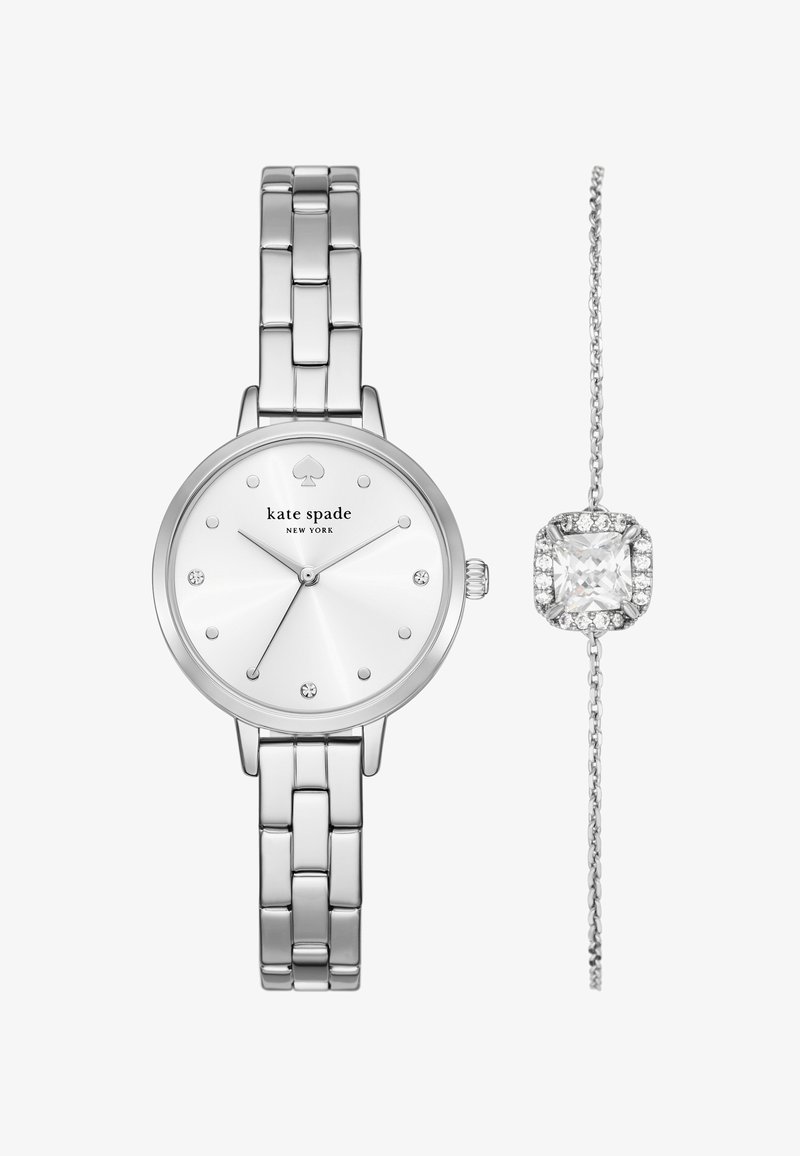 Srebrny zegarek z okrągłą tarczą, białym cyferblatem, diamentowymi akcentami i bransoletą ze stali nierdzewnej. Towarzyszy mu delikatna srebrna bransoletka z przezroczystym klejnotem.