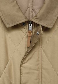 Giacca trapuntata beige con collo a camicia, chiusura a zip con tirante in pelle e bottoni a pressione in metallo. Tessuto testurizzato con cuciture diagonali.