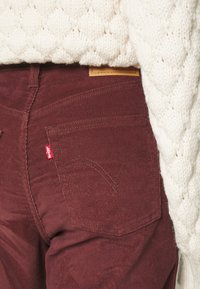 Bordowe spodnie z korduroyu z detalami na tylnej kieszeni, czerwoną metką Levi's i kontrastową, tan skórzaną łatą. Sweter ma grubą, teksturowaną dzianinę.