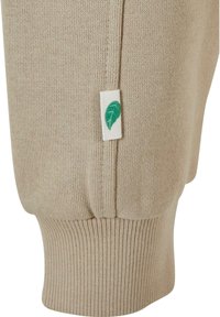 Beige strikkegenser med ribbestrikkede mansjetter, med et lite hvitt merke med et grønt bladdetalj. Myk tekstur og avslappet fasong.