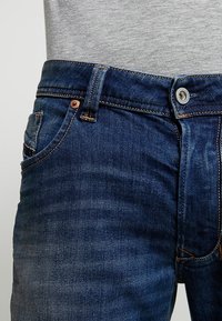 Närbild av mörkblå denimjeans midja och framficka, med synliga sömmar och kopparnippler, bar med en ljusgrå skjorta.