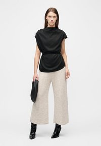 Femeie purtând un top negru fără mâneci cu o curea, pantaloni bej largi și scurți, botine negre din piele lacuită și ținând o poșetă clutch neagră.