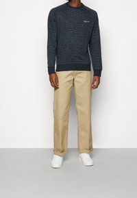 Maglione girocollo in tessuto navy con logo dorato, abbinato a pantaloni beige a gamba dritta e sneakers bianche. Design classico, vestibilità casual.