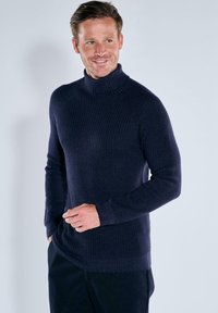 Lächelnder Mann mit kurzen Haaren, der einen navyblauen, gerippten Rollkragenpullover und dunkle Hosen trägt, vor einem einfarbigen hellen Hintergrund stehend.