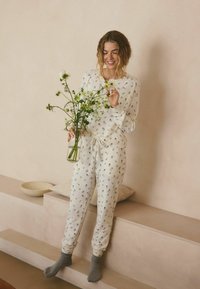 Helles Creme-Pyjama-Set mit gerippter Textur und floralem Muster. Das Modell hält eine Glasvase mit Grünpflanzen und weißen Blumen. Graue Socken.