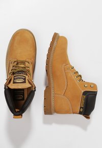 Botas de trabajo de cuero en color marrón claro con un diseño robusto, que presentan acentos en negro, herrajes en tono dorado, cordones redondos y una suela gruesa y estriada.