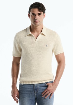 Junger Mann, der frontal schaut und ein cremefarbenes, kurzärmeliges Strick-Poloshirt sowie blaue Jeans trägt, mit einer Hand in der Hosentasche.