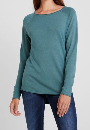Femme portant un pull à col rond manches longues couleur sarcelle, associé à un jean bleu foncé, debout devant un fond gris uni.