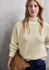 Maglione a coste crema con collo alto, abbinato a una borsa in pelle marrone, indossato su jeans scuri. Tessuto strutturato e vestibilità comoda.