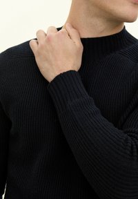 Svart stickad tröja med hög ribbad krage och texturerad yta. Närbild på en hand som justerar kragen.