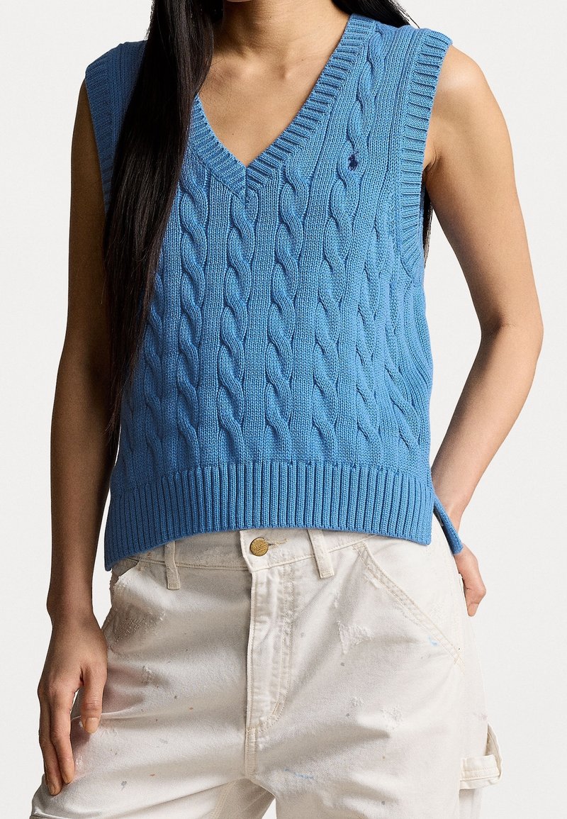 Himmelblaues, kabelgestricktes, ärmelloses Sweatshirt mit V-Ausschnitt, geripptem Saum und kleinem Logo auf der linken Seite, kombiniert mit cremefarbenen Hosen.