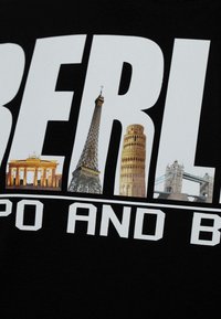 Schwarzes T-Shirt mit großem weißen Text und Bildern von ikonischen Sehenswürdigkeiten: Brandenburger Tor, Eiffelturm, Schiefe Turm von Pisa und Tower Bridge.
