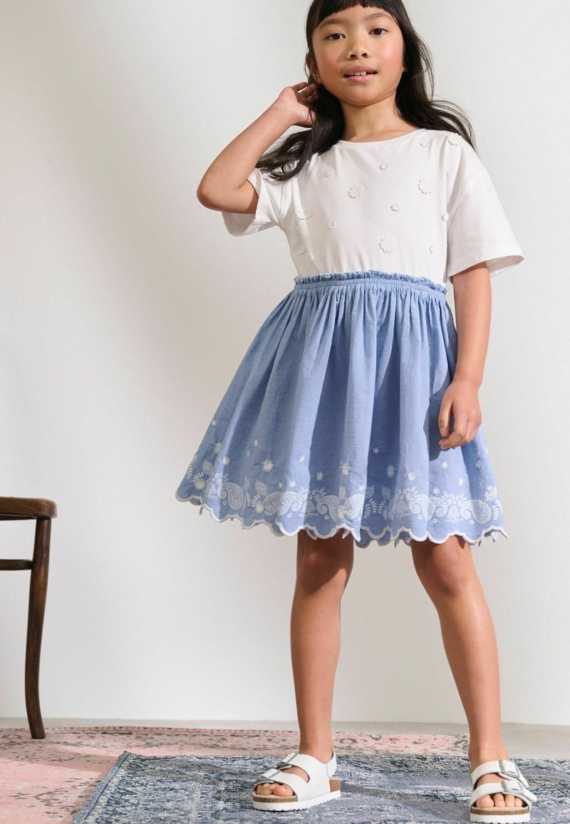Jeune fille debout, main près des cheveux, portant une chemise blanche avec des détails floraux, une jupe bleue avec une broderie blanche et des sandales blanches.