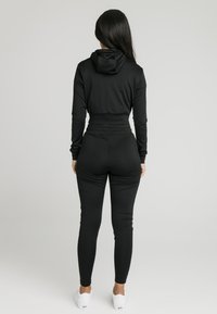 Musta urheilusetti, joka sisältää lyhyt mallisen hupullisen paidan ja kapeat housut. Sileä kangas, joustava vyötärönauha ja istuva malli lisäävät mukavuutta.