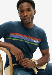 Camiseta de algodón azul marino con rayas horizontales en rojo, púrpura, amarillo y verde. Presenta un pequeño logo en el pecho. Corte casual.