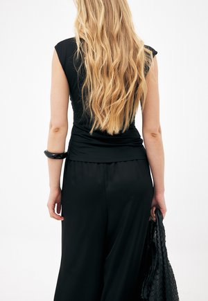 Vrouw met lang golvend blond haar, gekleed in een zwarte mouwloze top en brede zwarte broek, met in haar rechterhand een zwarte tas met textuur.