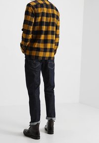 Persona che indossa una camicia a quadri nera e senape, jeans scuri arrotolati e stivali neri in pelle con lacci, in piedi su uno sfondo bianco.