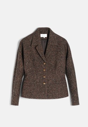Femme portant une veste ceinturée texturée marron foncé, une jupe midi assortie et des bottes marron foncé montant aux genoux, debout devant un fond uni.