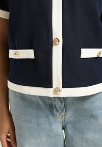 Top azul marino con ribete blanco, botones decorativos dorados, bolsillos falsos, llevado con vaqueros de mezclilla azul claro en una persona.