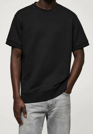 Mand iført en ensfarvet sort kortærmet sweatshirt og lysegrå jeans med den ene hånd i lommen mod en lys baggrund.