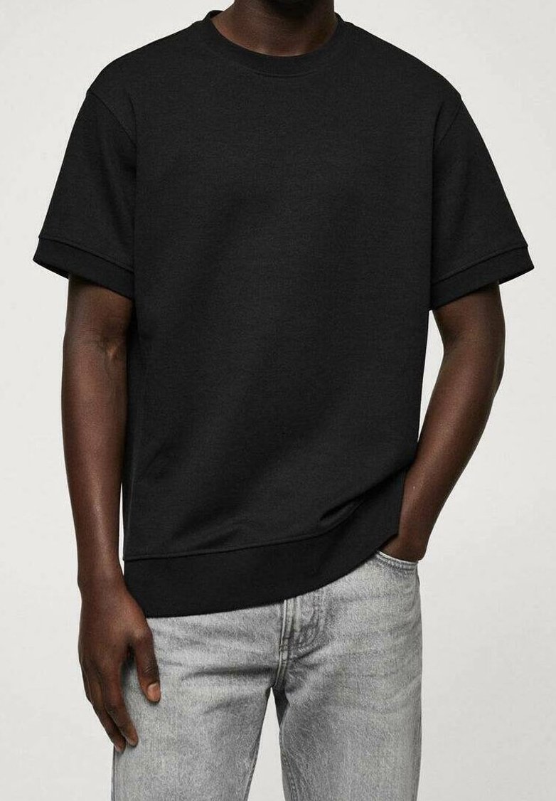 Homme portant un sweat-shirt noir uni à manches courtes et un jean gris clair, une main dans la poche, sur un fond clair.