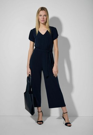 Jumpsuit bleu marine avec un décolleté en V, des manches courtes et une taille ceinturée, doté de jambes larges. Complété par des talons noirs à lanières et un sac fourre-tout sombre.
