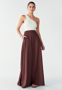 Vrouw in een witte off-shoulder ribgebreide top, lange bruine rok met zakken, gouden oorbellen, armbanden en zwarte sandalen.