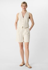 Beżowa kamizelka dopasowana z głębokim dekoltem w kształcie V i trzema guzikami, w połączeniu z pasującymi szortami z plisami. Ubranie wykonane jest z gładkiego, lekkiego materiału.