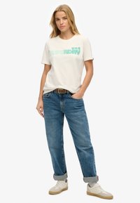 T-shirt z bawełny w kolorze off-white z zielonym napisem "SUPERDRY", połączony z niebieskimi dżinsami i kremowymi trampkami. Swobodny krój z podwiniętymi mankietami.