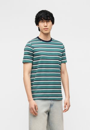 Ung mand iført grøn, marineblå og hvid stribet kortærmet skjorte og lyseblå jeans mod en ensfarvet hvid baggrund.