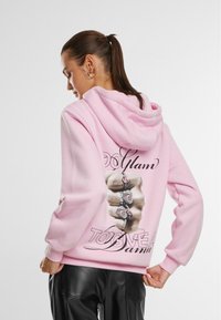 Roosa kapuutsiga sweatshirt, mille seljal on ringselt kaunistatud käe graafika ja stiilised tekstid. Tekstuur näeb välja pehme.