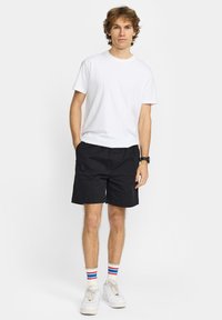 Junger Mann steht und trägt ein schlichtes weißes T-Shirt, schwarze Shorts, weiße Turnschuhe und weiße Socken mit roten und blauen Streifen.