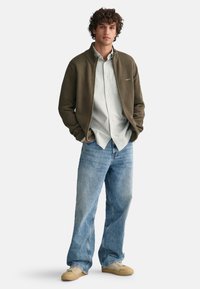 Giacca zip-up verde oliva sopra una camicia button-up a righe chiare, abbinata a jeans baggy blu chiaro e sneakers color sabbia.