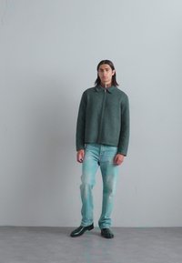Dunkelgrüne Wollmischung Jacke mit Kragen und durchgehendem Reißverschluss, kombiniert mit hellblauen Jeans und schwarzen Schuhen. Insgesamt glatte Textur.