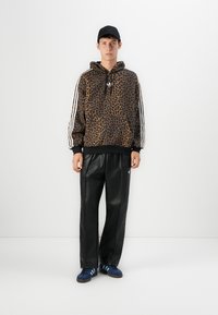 Uomo che indossa un felpa con stampa leopardata e strisce bianche, pantaloni di pelle nera, sneakers blu e cappellino nero, in piedi di fronte a un muro bianco.