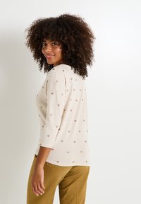 Femme aux cheveux bouclés portant un pull beige à petits motifs en forme de cœur et un pantalon moutarde, souriant et regardant par-dessus son épaule.