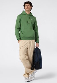 Felpa verde in cotone con cappuccio regolabile e tasca frontale; pantaloni cargo beige con tasche laterali; tiene uno zaino blu scuro.