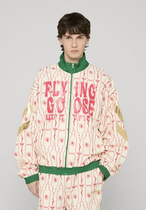 Young man wearing cream tracksuit with red bottle patterns and large red text, green collar and cuffs, and gold chevron stripes on sleeves.