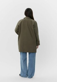 Giacca oversize di colore verde oliva con taglio dritto. Presenta maniche arrotolate e un dettaglio sul retro, abbinata a jeans a gamba larga di colore blu chiaro.